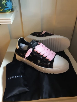 Numeris sneakers med rosa skosnören - Snygga sneakers från Numeris med svart och vit ovandel i textil och läderdetaljer. Skorna har breda rosa skosnören och en chunky vit sula. Perfekta för dig som vill sticka ut med en cool och modern look.
