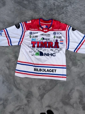 Timrå IK hockeytröja signerad - Säljer en vit och röd hockeytröja från Timrå IK, tillverkad av Bauer. Tröjan har blå och röda detaljer, flera sponsorer tryckta på framsidan och är signerad av spelare. Perfekt för dig som vill visa ditt stöd för laget eller samla på sportmemorabilia. Ps vissa autografer är från legendarer och vissa från nuvarande 
