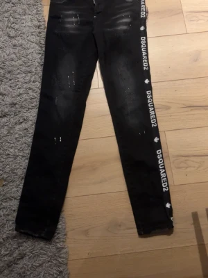 Svarta DSQUARED2 jeans med tryck - Säljer ett par svarta jeans från DSQUARED2 med vit logotyp längs sidorna och slitna detaljer framtill. Jeansen har rak passform och klassisk femficksdesign. Perfekta för dig som gillar streetstyle och vill sticka ut med en trendig look.