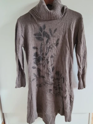Brun stickad polotröja med blommönster - Mysig brun stickad polotröja med svart blommönster framtill. Tröjan har långa ärmar, ribbade muddar och en hög, bred polokrage. Perfekt för kyliga dagar och enkel att styla med jeans eller leggings.