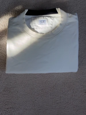 Vit sweatshirt från C.P. Company - Vit sweatshirt från C.P. Company med rund halsringning och ikonisk svart linsdetalj på ärmen. Tröjan har en clean look och är tillverkad i mjukt bomullsmaterial. Använd ett par gånger. 