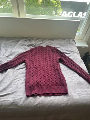 Vinröd kabelstickad tröja från GANT - Säljer en vinröd kabelstickad tröja från GANT i storlek S. Tröjan har rund halsringning, ribbade muddar och ett klassiskt flätmönster. Perfekt för dig som gillar stilrena och tidlösa plagg. Materialet är mjukt och skönt, troligtvis bomull.