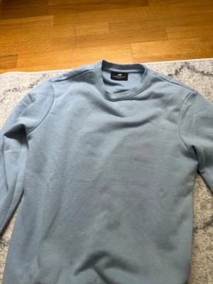 Ljusblå sweatshirt från H&M - En stilren ljusblå sweatshirt från H&M med rund halsringning och långa ärmar. Tröjan har en enkel design utan tryck eller mönster, vilket gör den lätt att matcha med olika outfits. Tillverkad i mjukt bomullsmaterial för en bekväm känsla.