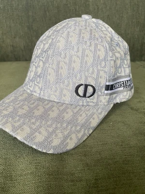 Beige mönstrad keps från Dior - Säljer en beige keps från Christian Dior med ikoniskt monogrammönster och broderad CD-logga framtill. Kepsen har justerbar rem bak och ett band med texten 'Christian Dior Paris' på sidan. Tillverkad i tyg med exklusiv känsla.