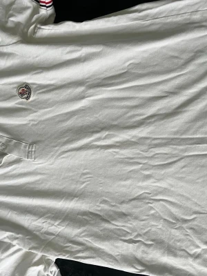 Vit pikétröja från Moncler - Klassisk vit pikétröja från Moncler med broderad logga på bröstet och knäppning vid halsen. Tröjan har krage med diskreta ränder i rött och blått. Perfekt till en clean och sportig stil.