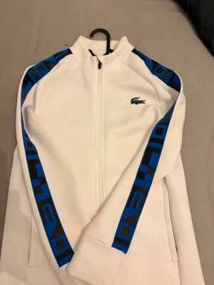 Vit hoodie från Lacoste Sport - Snygg vit hoodie från Lacoste Sport med hel dragkedja och blå detaljer längs ärmarna. Ikoniska krokodilloggan på bröstet och coolt mönster i blått och svart på ärmarna. Tillverkad i ett sportigt, ventilerande material som känns fräscht och modernt.