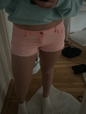 Ljusrosa jeansshorts med fransig kant - Säljer ett par ljusrosa jeansshorts med fransig kant nertill. Klassisk femficksmodell med knapp och dragkedja framtill. Perfekta för sommaren och ger en fräsch vibe till din outfit.