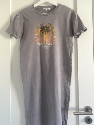 Grå t-shirtklänning från Garcia - T-shirtklänning i grått från Garcia med tryck av palm och texten 'Keep on the sunny side' i guld och orange. Rund halsringning, korta ärmar och rak passform. Perfekt för en avslappnad look.