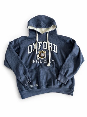 Blå Oxford University hoodie - Mörkblå hoodie med vit insida i huvan och vita dragsnören. Stor Oxford University-text och emblem broderat på bröstet. Klassisk känguruficka framtill och ribbade muddar. Perfekt till vardagen!