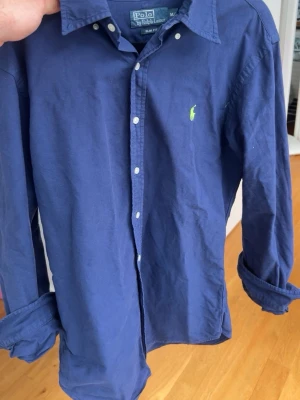 Blå skjorta från Polo Ralph Lauren - Klassisk blå skjorta från Polo Ralph Lauren med lång ärm och knappar framtill. Skjortan har den ikoniska gröna logotypen broderad på bröstet och är tillverkad i mjuk bomull. Perfekt till jeans eller chinos för en clean look.