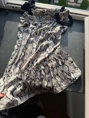 Medellång klänning med ormmönster i silke - Säljer en kort klänning med trendigt ormmönster i beige, svart och grå toner. Klänningen har volang nertill och svarta rosetter på axelbanden för en extra snygg detalj. Perfekt för dig som vill sticka ut med en klänning som min familj tillverkat, The Airen.