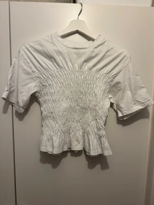 Vit smockad topp med kort ärm - Säljer en vit topp med smockad midja och korta ärmar. Toppen har en rund halsringning och lätt volang nedtill för en söt look. 