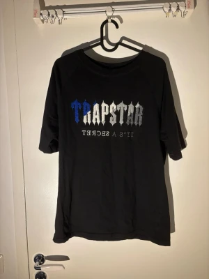 Trapstar t-shirt - Svart t-shirt från Trapstar med stort tryck i blått, vitt och grått på bröstet där det står 'TRAPSTAR IT'S A SECRET'. Klassisk rund hals och korta ärmar. Tillverkad i mjuk bomull för en bekväm känsla.