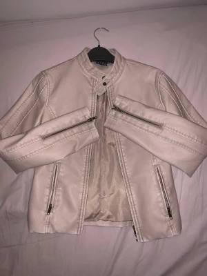 Ljusbeige skinnjacka från MINAIDE - Snygg ljusbeige skinnjacka från MINAIDE med ståkrage och dekorativa sömmar. Jackan har dragkedjor på ärmarna och framfickor samt en knappdetalj vid halsen. Perfekt för dig som vill ha en stilren och trendig look. Har aldrig använts innan är helt ny! 