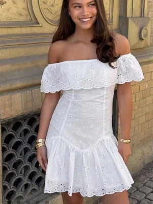 Vit offshoulder klänning med volang - Superfin vit kortklänning med offshoulder och volangkant. Klänningen har broderade blommor och är tillverkad i bomull, vilket gör den luftig och perfekt för varma dagar. Söt och feminin modell som ger en romantisk vibe.
