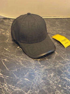 Svart keps från Fendi med FF-mönster - Snygg svart keps från Fendi med det klassiska FF-logomönstret ton-i-ton över hela kepsen. Böjd skärm och justerbar baksida. Tillverkad i ett slitstarkt tyg som ger en exklusiv känsla. Perfekt accessoar för att lyfta din streetwear-look.