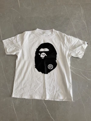 Vit BAPE t-shirt med ape-tryck - Säljer en vit t-shirt från BAPE med det ikoniska svarta ape-huvudet tryckt stort på både fram- och baksidan. Klassisk rund hals och korta ärmar. Perfekt för dig som gillar streetwear och vill sticka ut med en clean men ändå bold look.