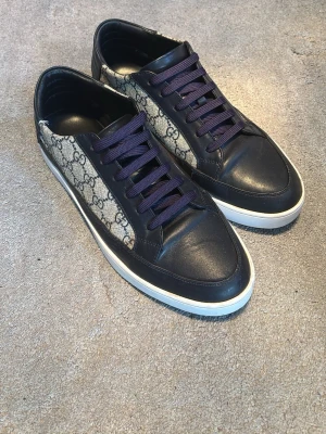 Gucci sneakers - Snygga sneakers från Gucci med klassiskt GG-monogram i beige och mörkblått. Skorna har mörkblå läderdetaljer, vita sulor och mörka snören. Perfekt för dig som vill ha en lyxig och trendig look med premiumkänsla.