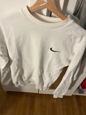Vit sweatshirt från Nike - Säljer en stilren vit sweatshirt från Nike med svart broderad Swoosh-logga på bröstet. Tröjan har rund halsringning, långa ärmar och ribbade muddar vid ärmslut och nederkant. Perfekt för en clean och sportig look.