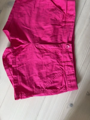 Rosa shorts i bomull - Säljer ett par snygga rosa shorts i bomull med normal passform och låg midja. Shortsen har knappstängning framtill och två fickor på sidorna. Perfekta för varma dagar och enkel att matcha med olika toppar. Inga fickor där bak vilket gör shortsen mer unika och coola. Svala och luftiga som passar bra till varmare sommardagar.