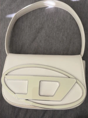 Vit Diesel handväska med metallogo - Diesel handväska. Har använt den 1-2 ggr. Har spår av smink på insidan som går att få bort med sminkborttagning. Vet ej om den är äkta eller inte.