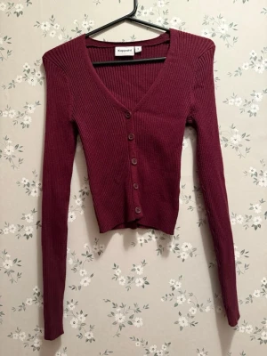 Vinröd ribbad kofta från Kappahl - Snygg vinröd kofta från Kappahl med ribbad struktur och v-ringning. Koftan har långa ärmar och knäpps med fem knappar framtill. Perfekt att slänga över en topp för en trendig lager-på-lager-look. Kanin finns i hemmet. I storleken s men är väldigt liten i storleken.