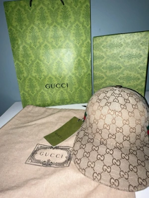 Beige keps från Gucci med GG-mönster - Säljer en beige keps från Gucci med det klassiska GG-monogrammet över hela kepsen. Kepsen har böjd skärm och detaljer i grönt och rött på sidan. Tillverkad i canvasmaterial och levereras med originalpåse och tagg.