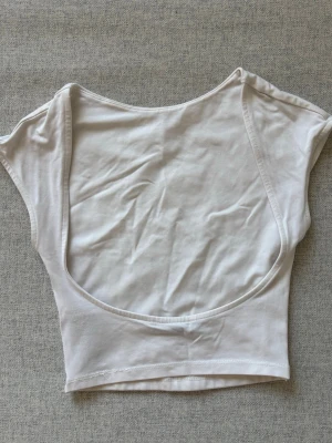 Vit croppad topp med öppen rygg - Säljer en vit croppad topp i mjuk bomull med korta ärmar och en stor, öppen rygg. Perfekt till höga jeans eller kjol för en trendig look. Enkel och stilren design utan mönster eller detaljer. Lite bikinimaterial fast tjockare. I storlek XS från Bershka.