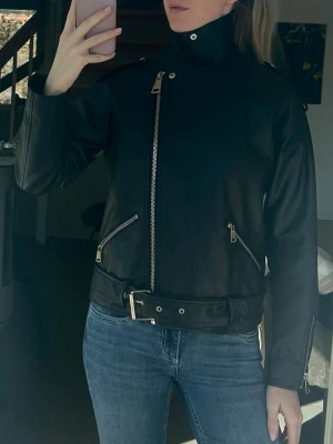 Svart skinnjacka med bälte - Snygg svart skinnjacka med hög krage, sned dragkedja och bälte i midjan. Jackan har två snedställda fickor med dragkedja och dragkedjor vid ärmsluten. Perfekt för dig som gillar en klassisk bikerstil.
