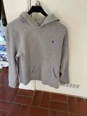 Grå hoodie från Polo Ralph Lauren - Klassisk grå hoodie från Polo Ralph Lauren med broderad marinblå logga på bröstet. Hoodien har huva, känguruficka och ribbade muddar vid ärmar och nederkant. Tillverkad i mjukt sweatshirtmaterial som är perfekt för en avslappnad stil.