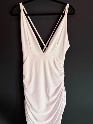 Vit omlottklänning med djup v-ringning - Snygg vit klänning med djup v-ringning och omlott-design både fram och bak. Klänningen har smala axelband som korsas i ryggen och en draperad effekt på sidan. Perfekt för dig som vill ha en stilren och trendig look.