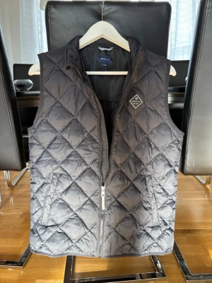 Svart dunväst från Gant - Snygg svart dunväst från Gant med quiltat mönster och hög krage. Västen har dragkedja framtill, två sidofickor och en diskret logotyp på bröstet. Perfekt att slänga över en hoodie eller tröja för extra värme och stil.