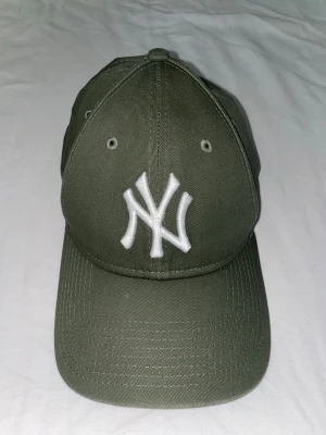 Olivgrön New Era NY keps - Snygg olivgrön keps från New Era med klassisk böjd skärm och broderad vit NY-logga framtill. Kepsen har ventilationshål och justerbar passform. Tillverkad i slitstarkt bomullstyg, perfekt för en sportig och trendig look.