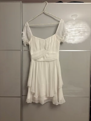 Kort vit klänning med volang  - Söt vit kortklänning med omlottdetalj och volangkant. Klänningen har puffiga kortärmar i transparent tyg och ett lager på lager-fall i kjolen. Perfekt för skolavslutningar eller studenten✨💞💝(klänningen har några slitningar som syns på bilderna, men inget som märks av i verkligheten) 