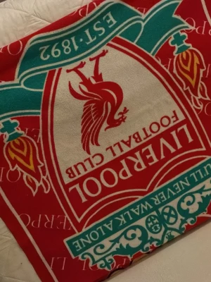Liverpool FC röd halsduk - Snygg röd halsduk från Liverpool FC med klubbens klassiska logga och texten 'You'll Never Walk Alone' i vitt och turkos. Perfekt accessoar för alla fans som vill visa sitt stöd på matchdagar eller i vardagen.