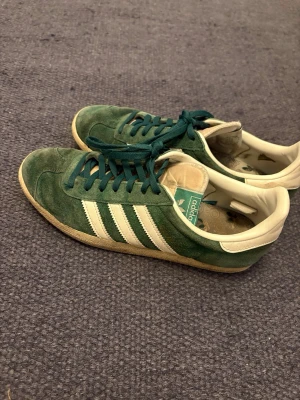 Gröna Adidas Gazelle sneakers - Snygga gröna Adidas Gazelle sneakers i mocka med klassiska vita ränder på sidorna och vit häl. Skorna har gröna skosnören och en gummisula med hexagonmönster. Perfekta för dig som gillar snygga sneakers till vardags. Passar 40 och 41.