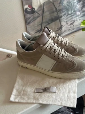Valentino Flycrews 41 - Säljer ett par snygga Valentino Garavani sneakers i beige mocka med vita detaljer och klassisk snörning. Skorna har en vit sula och diskreta nitar på hälen. Perfekta för dig som vill ha en stilren och exklusiv look. De är inte i det bästa skicket, därav är de lite billigare, saknar nitar, defekter i sulan och mockan, annars är de riktigt snygga 