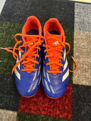 Adidas Predator blå fotbollsskor - Säljer ett par Adidas Predator fotbollsskor i blått med vita detaljer och orange snörning samt innersida. Skorna har klassisk låg profil och är tillverkade i syntetmaterial för bra bollkontroll. Perfekta för dig som vill sticka ut på planen.
