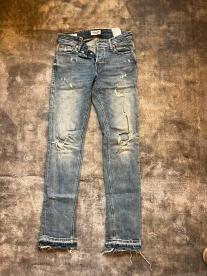 Blå slitna jeans från Nudie Jeans - Säljer ett par blå jeans från Nudie Jeans med slitningar och hål på benen för en riktigt trendig look. Modellen är rak men har sytts ut se bild till bootcut  med råa, fransiga benslut och klassisk femficksdesign. Perfekta för dig som gillar en avslappnad och edgy stil. Skulle säga att dem är små i storleken🥰