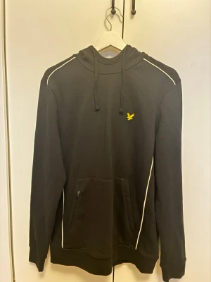 Svart (OANVÄND) hoodie från Lyle & Scott - Svart hoodie från Lyle & Scott med vit piping längs axlar och sidor. Klassisk huva med dragsko, magficka med dragkedja och gul logga på bröstet. Tillverkad i mjuk bomullsblandning, perfekt för en chill och stilren look. Tröjan är helt oanvänd.