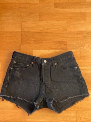 Svarta Levi's 501 jeansshorts med rå kant W30 - Säljer ett par svarta jeansshorts från Levi's 501 med råa, fransiga kanter och klassisk femficksdesign. Storlek W30. Shortsen har hög midja och knappgylf. Perfekta för sommaren och har en avslappnad, vintage look.