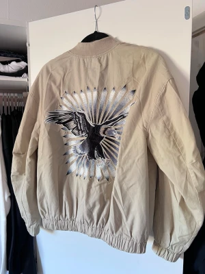 Beige y2k jacka med örn-tryck på baksidan, H&M - stl S - Tynnre beige y2k bomberjacka från H&M med snyggt broderi av en örn på ryggen. Säljer pga att den är lite för liten för mig.  Accepterar alla bud!
