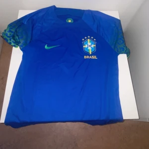 MarinBlå Brasilien Nike fotbollströja  - Snygg blå fotbollströja från Nike med Brasilien-emblem och fem stjärnor på bröstet. Tröjan har gröna detaljer på ärmarna och ett tryckt Nike-märke. Perfekt för dig som älskar fotboll och vill stötta Brasilien med stil. Pris kan diskuteras 🤝