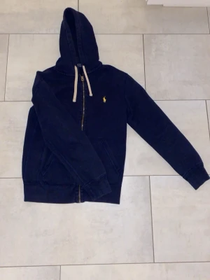 Hoodie från Ralph Lauren (kom med bud!) - Mörkblå hoodie med dragkedja från Polo Ralph Lauren. Klassisk gul logga på bröstet, vita snören i huvan och två fickor framtill. Tillverkad i mjuk bomullsmix som är skön att bära. Perfekt för en avslappnad och stilren look. Kom med bud!
