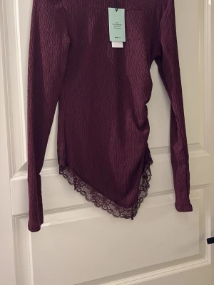 Vinröd topp med spetskant från Gina Tricot - Vinröd långärmad topp från Gina Tricot med asymmetrisk nederkant och dekorativ spetskant. Materialet är lätt skrynklat och toppen har en slim passform som framhäver siluetten. Perfekt för dig som gillar unika detaljer och en trendig look.