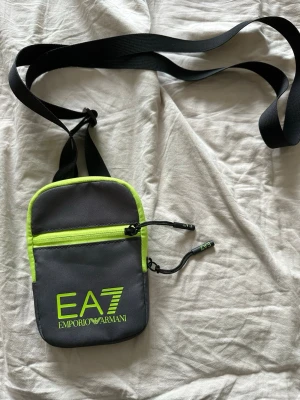 EA7 Emporio Armani axelväska grå/neon - Snygg axelväska från EA7 Emporio Armani i grått med neonlime detaljer och logga framtill. Väskan har justerbar svart axelrem, dragkedja upptill och en extra ficka med dragkedja på framsidan. Tillverkad i slitstarkt syntetmaterial.