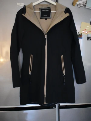 Svart regnjacka från Ilse Jacobsen - Snygg svart regnjacka från Ilse Jacobsen med beige insida och dragkedjor i kontrastfärg. Jackan har huva, långa ärmar och två praktiska fickor med dragkedja framtill. Perfekt för regniga dagar och enkel att matcha med olika outfits.