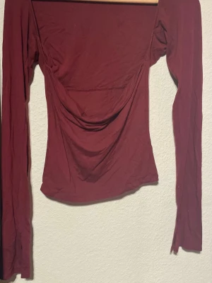 Vinröd långärmad topp - Snygg vinröd långärmad topp med draperad front och bred urringning. Toppen har en tight passform och är tillverkad i mjukt stretchigt material, perfekt för att lyfta din outfit. Passar dig som gillar trendiga och stilrena plagg.