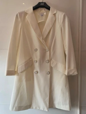Vit dubbelknäppt klänning/blazer som ny. - Stilren vit klänning/blazer med dubbelknäppning och breda slag. Kappan har två stora fickor framtill och är tillverkad i ett slätt tyg som ger en elegant look. Perfekt för dig som vill ha en klassisk och tidlös stil. Använd en gång.