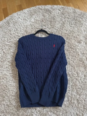 Mörkblå kabelstickad tröja Polo Ralph Lauren - Kabelstickad mörkblå tröja från Polo Ralph Lauren med rund halsringning och röd broderad logga på bröstet. Tröjan har långa ärmar och ribbade muddar vid ärmslut och nederkant. Perfekt för lager på lager och klassisk stil.